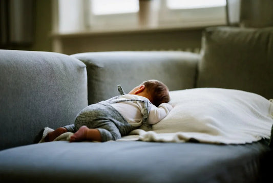 bébé qui dort sur son ventre sur un oreiller posé sur un canapé gris de salon